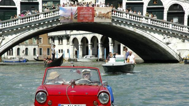 Ein rotes Amphicar fährt durch die Kanäle von Venedig, im Hintergrund eine Brücke mit vielen Zuschauern.