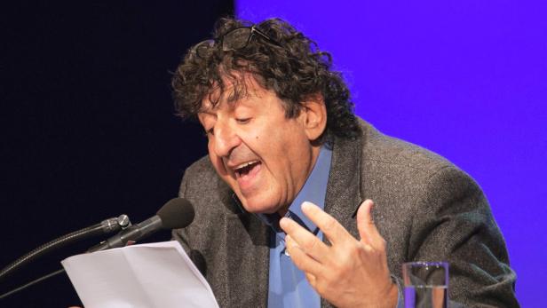 Ein Mann mit lockigem Haar liest von einem Blatt Papier vor einem Mikrofon.