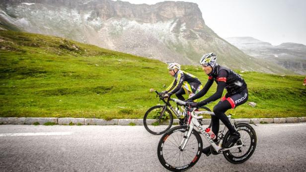 Zwei Radfahrer fahren eine asphaltierte Straße entlang, im Hintergrund grüne Hügel und Berge.