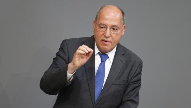 Ein Mann mit Brille und Anzug gestikuliert vor einem grauen Hintergrund.