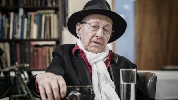 Ein älterer Mann mit Hut und Brille sitzt mit einer Kamera an einem Tisch.