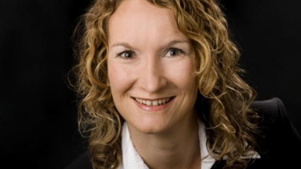 Eine lächelnde Frau mit blonden, lockigen Haaren und einem schwarzen Blazer vor einem schwarzen Hintergrund.