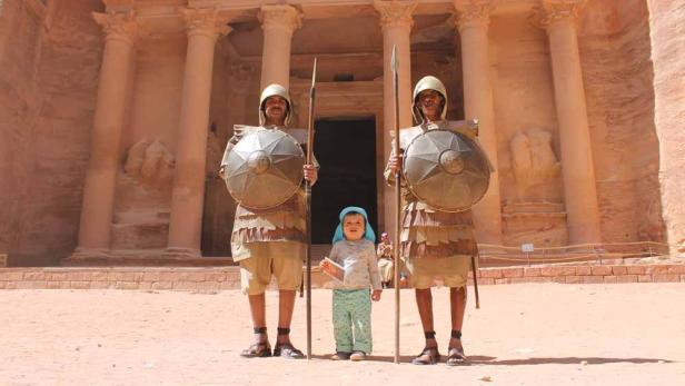Zwei Männer in Kostümen mit Schilden und Speeren stehen neben einem kleinen Kind vor dem Schatzhaus in Petra.