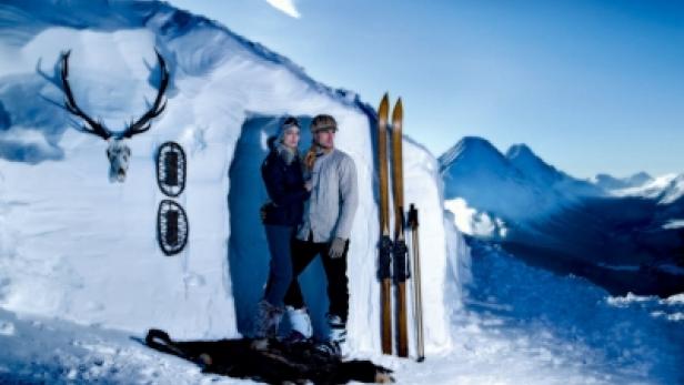 Ein Paar steht vor einem Iglu in einer verschneiten Berglandschaft.