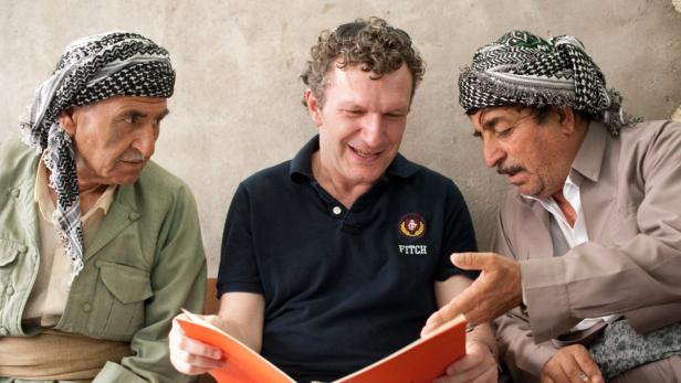 Zwei ältere Männer mit traditionellen Kopfbedeckungen betrachten mit einem Mann ein Buch.