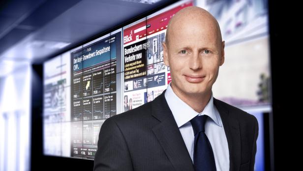 Ein Mann im Anzug steht vor einer Wand aus Bildschirmen mit Nachrichten und Grafiken.