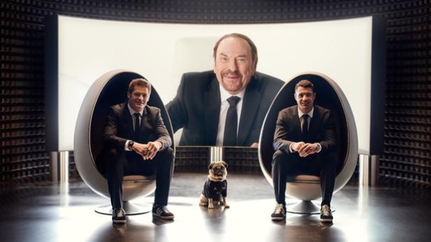 Zwei Männer in Anzügen sitzen in futuristischen Stühlen, während ein Mann auf einem Bildschirm und ein Mops zusehen.