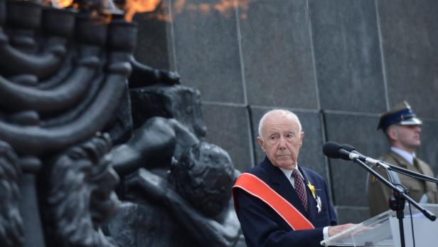 Ein älterer Mann mit einer Schärpe steht an einem Rednerpult vor einem Denkmal.