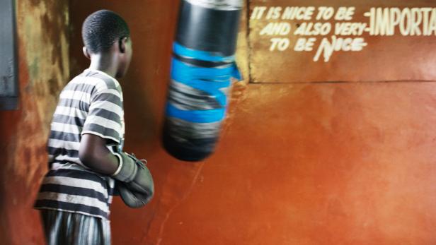 Ein junger Boxer mit Boxhandschuhen steht vor einem Boxsack in einem Raum mit einem Spruch an der Wand.