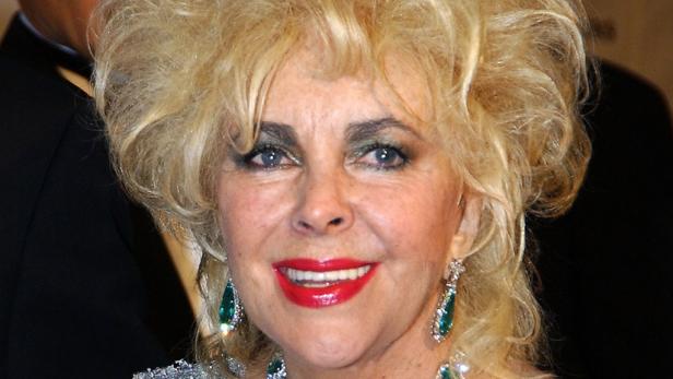 Elizabeth Taylor mit blonden Locken, smaragdgrüner Halskette und passenden Ohrringen lächelt.
