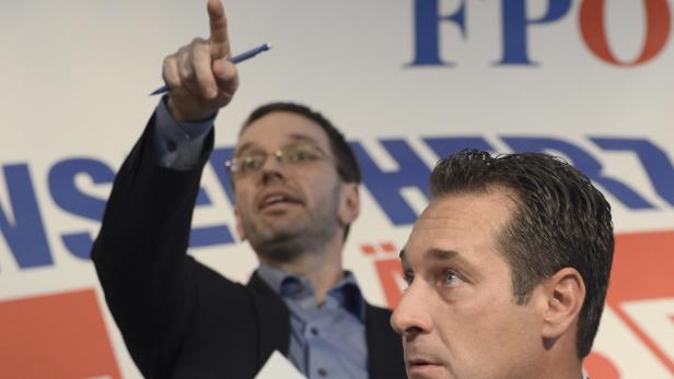 Zwei Männer stehen vor einem FPÖ-Logo, einer zeigt mit einem Stift nach oben.