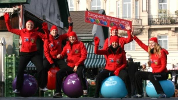 Eine Gruppe von Frauen in roten Jacken sitzt auf Hüpfbällen und hält ein Schild mit der Aufschrift hopperinvasion.com.