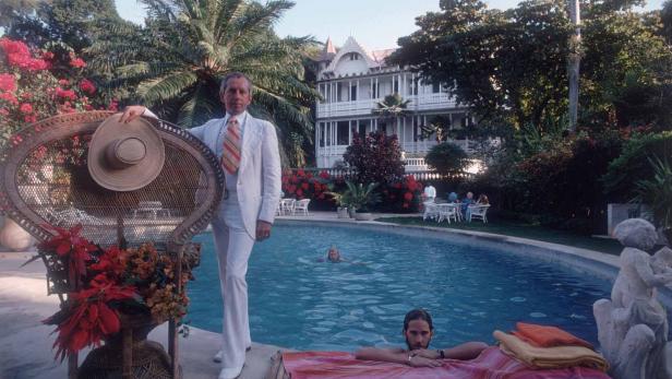 Ein Mann im Anzug posiert neben einem Pool vor einem weißen Haus.
