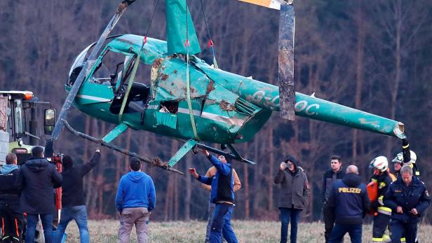 Ein beschädigter, grüner Hubschrauber wird von einem Kran über ein Feld gehoben. Polizisten und Feuerwehrleute stehen daneben.