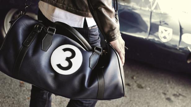 Eine Person mit einer blauen Reisetasche mit der Nummer 3 vor einem Oldtimer.
