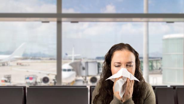Kranke Frau schnäuzt sich die Nase mit Flugzeug im Hintergrund.