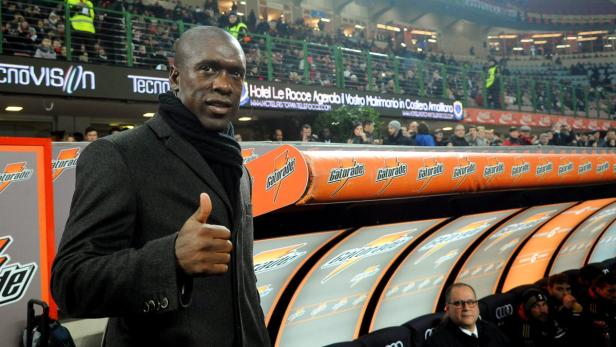 Ein Mann im Mantel steht vor der Auswechselbank in einem Fußballstadion und zeigt den Daumen nach oben.