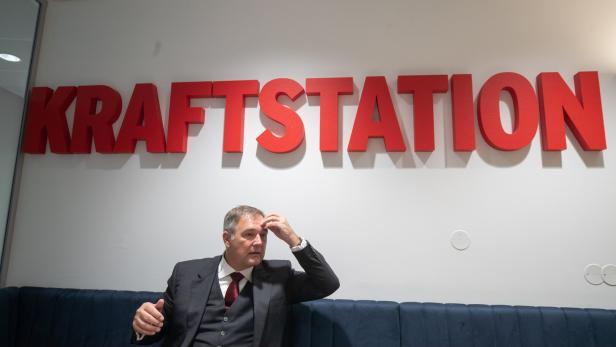 Ein Mann im Anzug sitzt vor einem großen roten „KRAFTSTATION“-Schriftzug und hält sich die Stirn.
