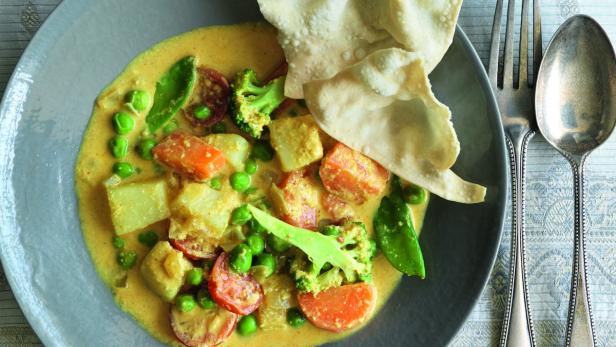 Ein Teller mit Gemüsecurry und Papadams, garniert mit Erbsen, Brokkoli und Karotten.