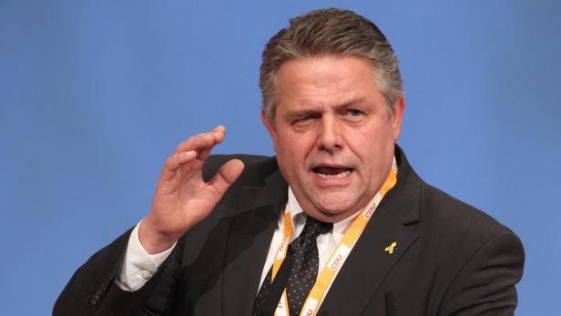 Ein Mann in einem Anzug gestikuliert vor einem blauen Hintergrund.