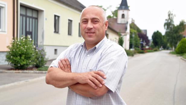 Ein Mann steht mit verschränkten Armen auf einer Straße in einer Ortschaft.