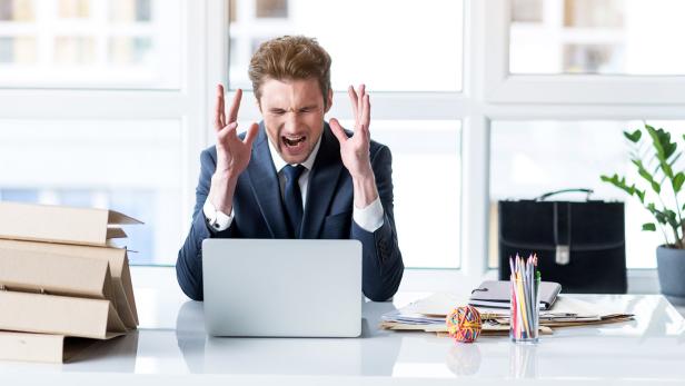 Ein frustrierter Mann im Anzug schreit vor seinem Laptop am Schreibtisch.