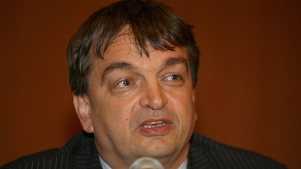 Ein Mann spricht in ein Mikrofon vor einem orangefarbenen Hintergrund.
