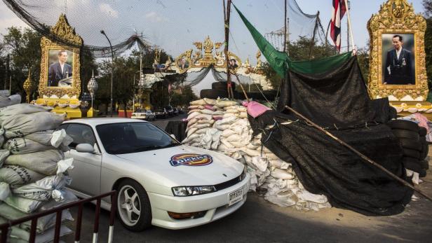Ein weißes Auto mit Dickies-Logo steht vor Sandsäcken und Porträts des thailändischen Königs.