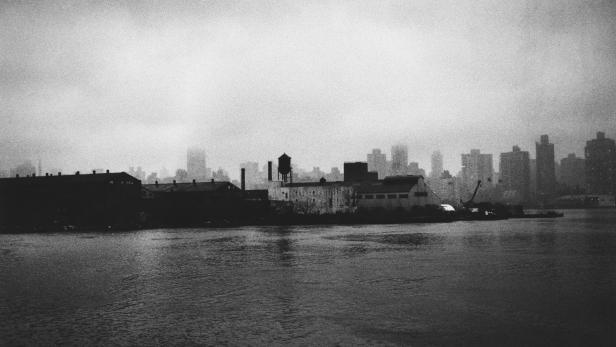 Eine Schwarzweißaufnahme einer Stadtlandschaft am Wasser unter einem trüben Himmel.