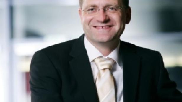 Ein lächelnder Mann mit Brille, Anzug und Krawatte vor einem Fenster.