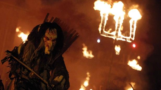 Ein maskierter Krampus mit Rute steht vor einem Feuer.