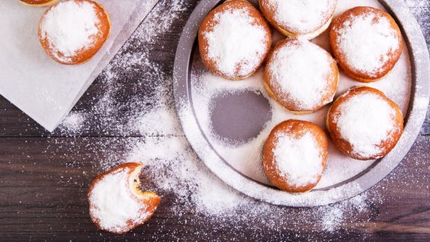 Mehrere mit Puderzucker bestäubte Krapfen auf einem Backblech und einem Küchentuch.