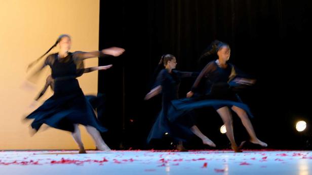 Mehrere Tänzerinnen in schwarzen Kleidern führen eine Performance auf einer Bühne mit roten Blütenblättern auf.