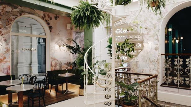 Eine weiße Wendeltreppe in einem hellen Raum mit vielen Pflanzen und dekorativen Wandmalereien.