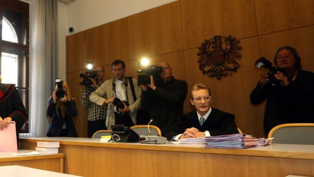 Ein Richter sitzt vor einem Stapel Akten, während Fotografen ihn mit Blitzlicht fotografieren.