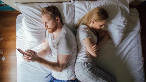Young couple lying in bed back to back, man using phone and woman frowning