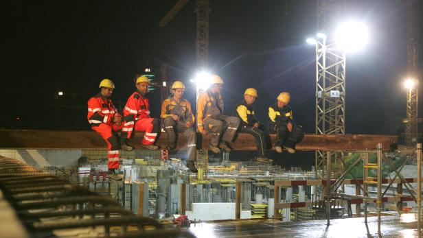 Mehrere Bauarbeiter sitzen nachts auf einem Stahlträger vor einer Baustelle.