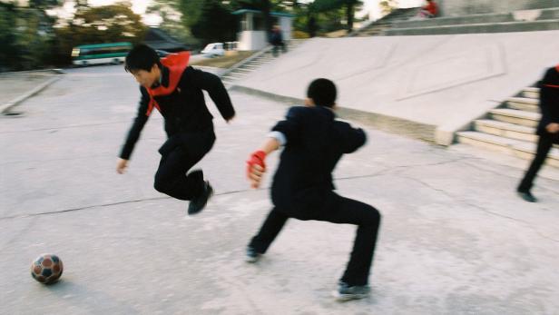 Zwei Jungen in Anzügen spielen mit einem Fußball auf einem Platz.