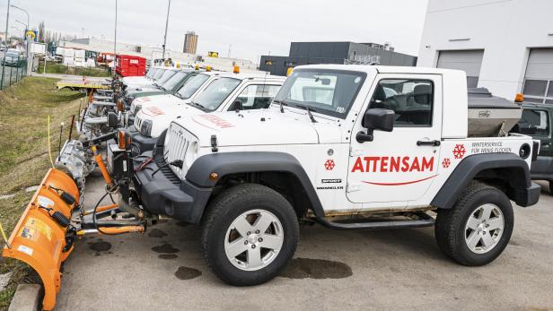 Mehrere weiße Fahrzeuge mit Schneepflügen und orangefarbenen Warnleuchten stehen nebeneinander auf einem Parkplatz.
