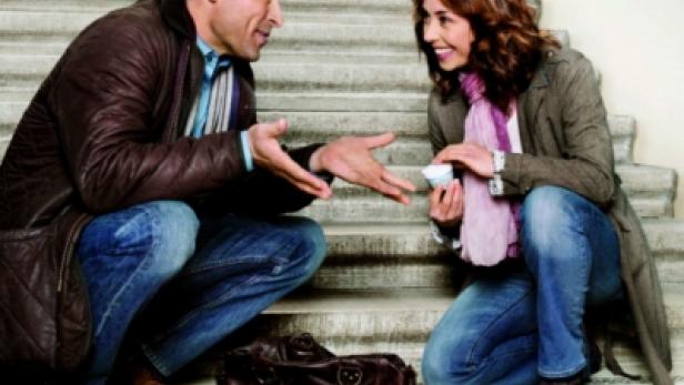 Ein Mann und eine Frau knien auf einer Treppe, während Gegenstände aus einer Handtasche gefallen sind.