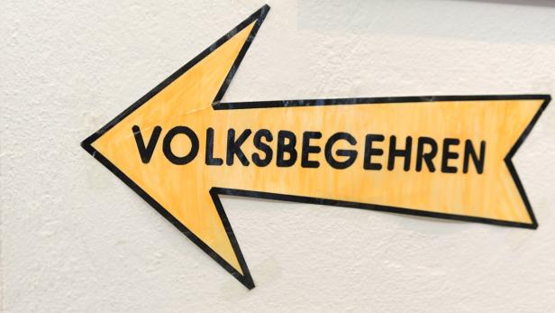 Ein gelber Pfeil an einer Wand zeigt nach links und trägt die Aufschrift „Volksbegehren“.