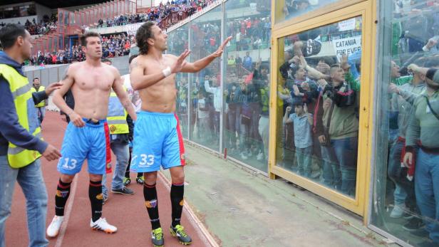Zwei Fußballspieler jubeln den Fans hinter einer Glaswand zu.
