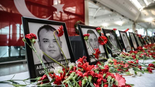 Auf einem Tisch stehen gerahmte Porträts, geschmückt mit roten und weißen Blumen, vor einer türkischen Flagge.