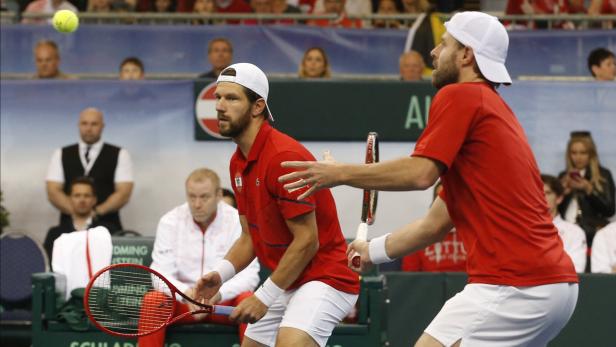 Zwei Tennisspieler in roten Trikots und weißen Kappen auf einem Tennisplatz.