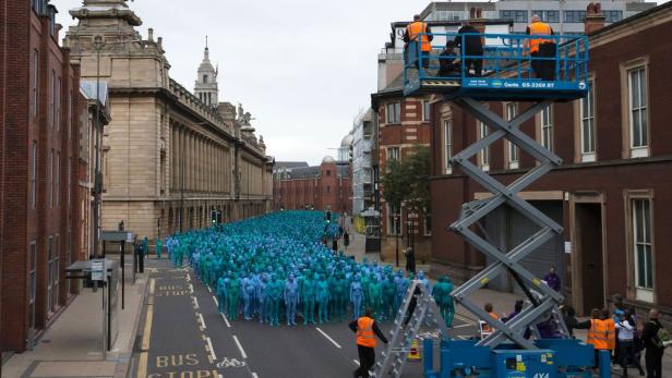 Eine große Gruppe blau bemalter Menschen steht auf einer Straße, die von Gebäuden gesäumt ist.