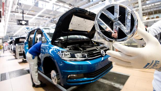 Ein Mitarbeiter zeigt ein VW Logo kurz vor Einbau in einen Volkswagen Touran in der Endmontage