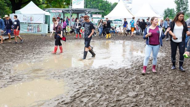 Menschen mit Gummistiefeln waten durch den Schlamm auf einem Festivalgelände.