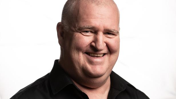 Ein lächelnder Mann mit Glatze und schwarzem Hemd vor einem weißen Hintergrund.