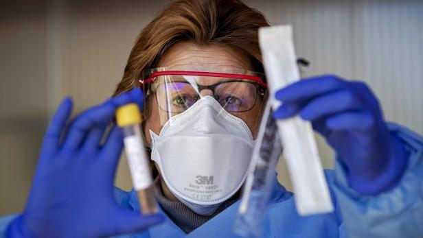 Eine medizinische Fachkraft mit Schutzkleidung hält ein Teströhrchen und einen Tupfer.