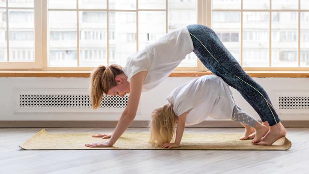 Young mother doing yoga with 3-years girl in front of window. Downward facing dog asana